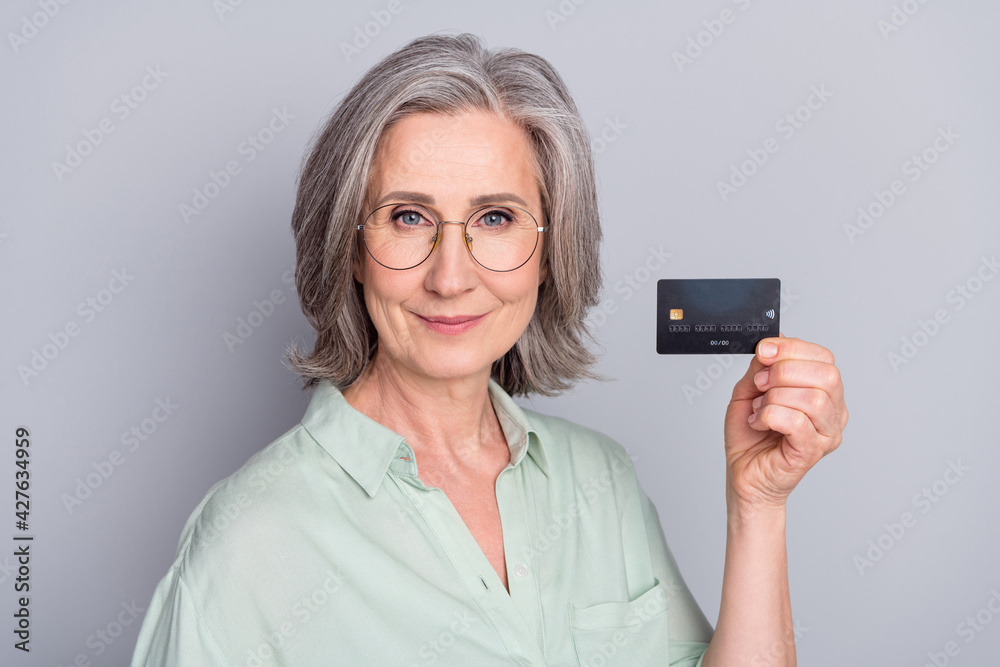 Photo of mature smiling beautiful woman in glasses showing credit card savings salary isolated on grey color background Photo of mature smiling beautiful woman in glasses showing credit card savings salary isolated on grey color background