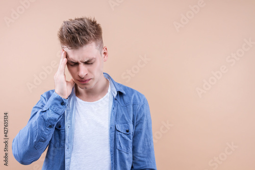 Young handsome man wearing denim shirt standing over isolated beige background tired rubbing, touching head feeling fatigue and headache. Stress and frustration concept. Cold flu virus symptoms.