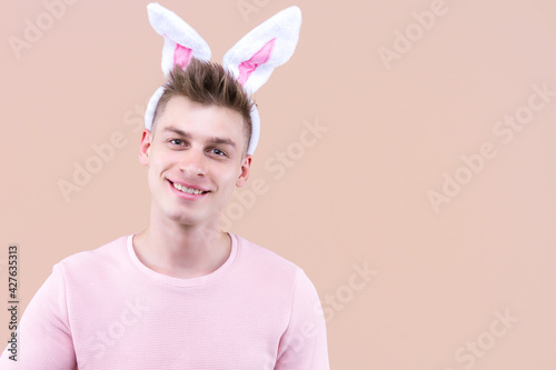 Young handsome man wearing traditional bunny rabbit ears is standing over isolated beige background. Funny male is looking with smile on face, natural expression. Happy Easter and spring concept.