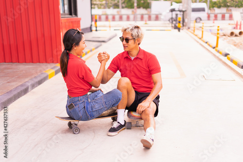 Asian handsome man and beautiful woman sit on skateboards  catch hands at skateboard way in daylight time  summer holiday