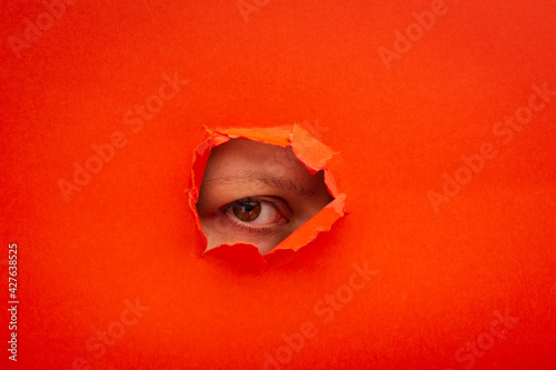 Red color paper with a ripped hole and watching eye from it, the concept of rumors and peeping. Funny picture of a woman's eye looking for interesting projects.