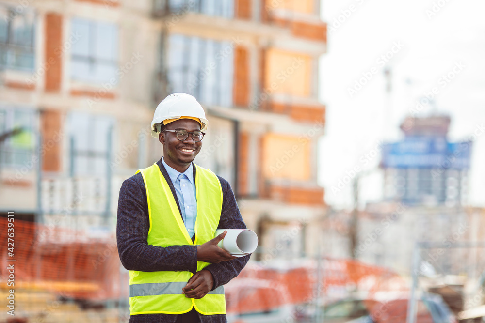 Portrait of African American man architect wearing a vest and helmet, he stands and holding a blueprints with the sky and urban in background. Engineer and architect concept.