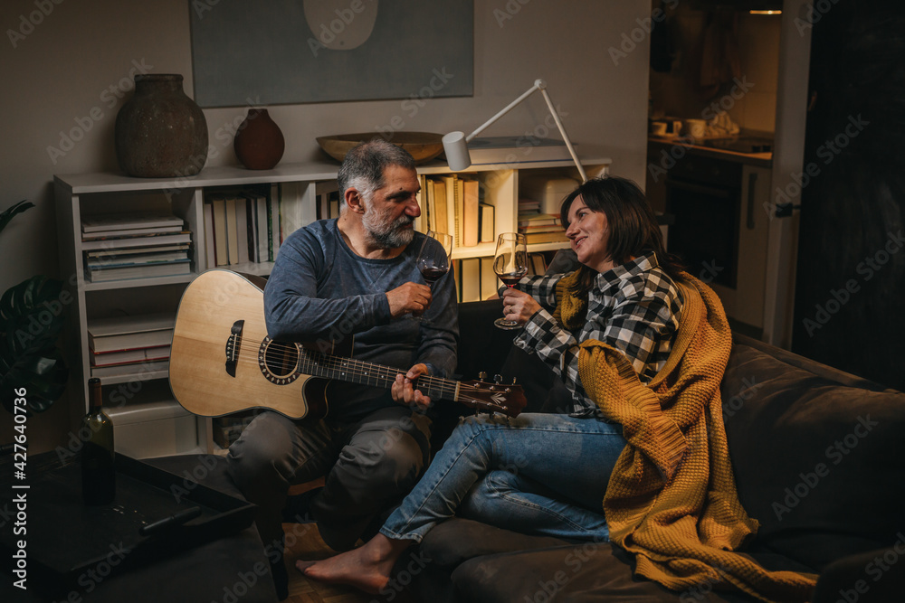 middle age romantic couple drinking wine while sitting on sofa at home. Man playing acoustic guitar