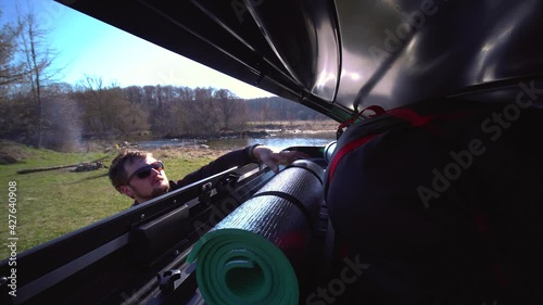 A man puts things in the roof rack of a car or in a cargo box, on a blurred background of a river and trees, on a summer day. family trip. Close-up Inside view.