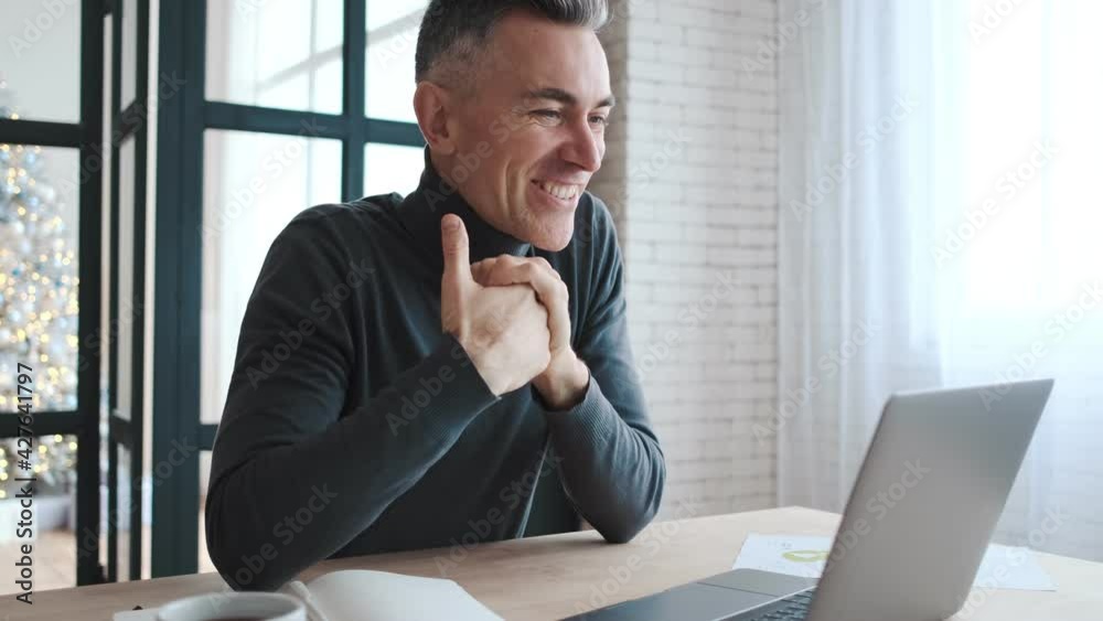 A side view of a mature smiling man sitting at the desk in front of the monitor while talking over a video call in the kitchen A side view of a mature smiling man sitting at the desk in front of the monitor while talking over a video call in the kitchen