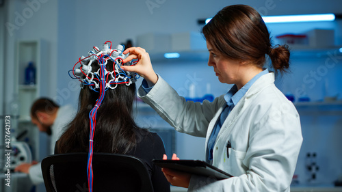 Woman patient wearing performant eeg headset scanning brain electrical activity in neurological research laboratory while medical researcher adjusting it, examining nervous system typing on tablet. photo