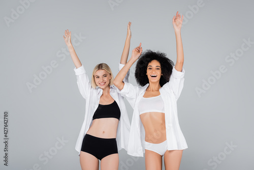Fototapeta Naklejka Na Ścianę i Meble -  excited multiethnic women in underwear and white shirts posing with raised hands isolated on grey