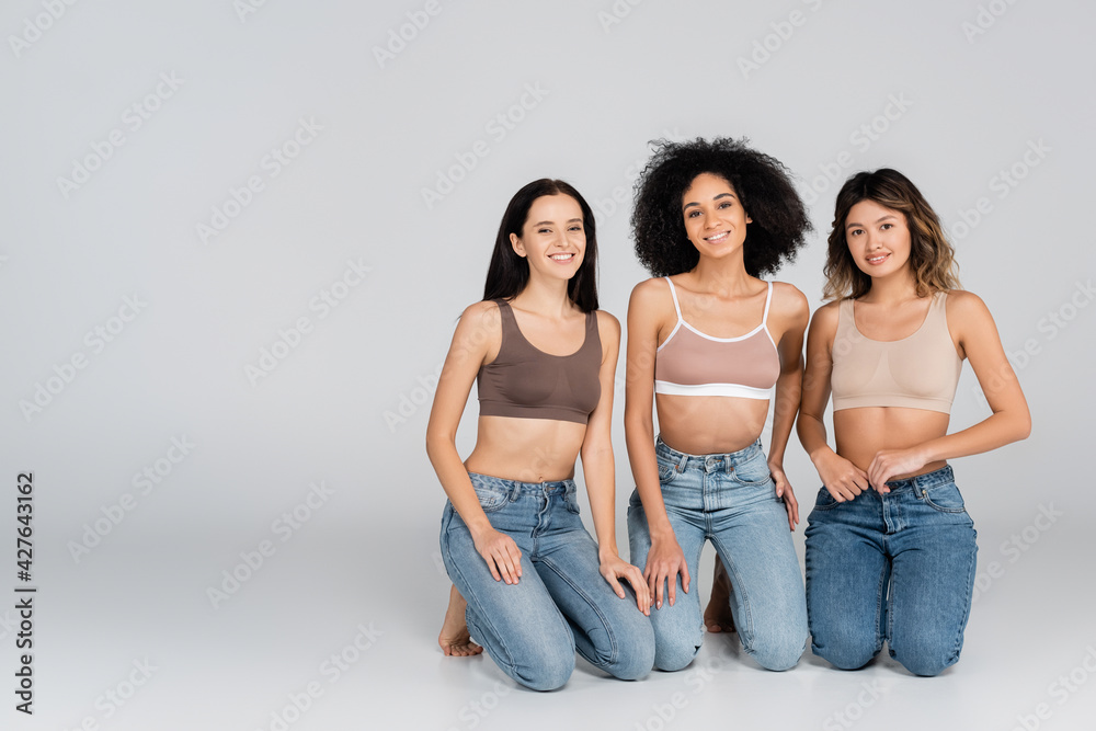 happy interracial women in bras and jeans looking at camera on grey happy interracial women in bras and jeans looking at camera on grey
