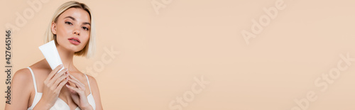 blonde woman looking at camera while holding cosmetic cream isolated on beige, banner