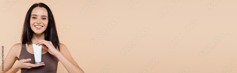 young woman smiling at camera while holding cosmetic cream isolated on beige, banner young woman smiling at camera while holding cosmetic cream isolated on beige, banner