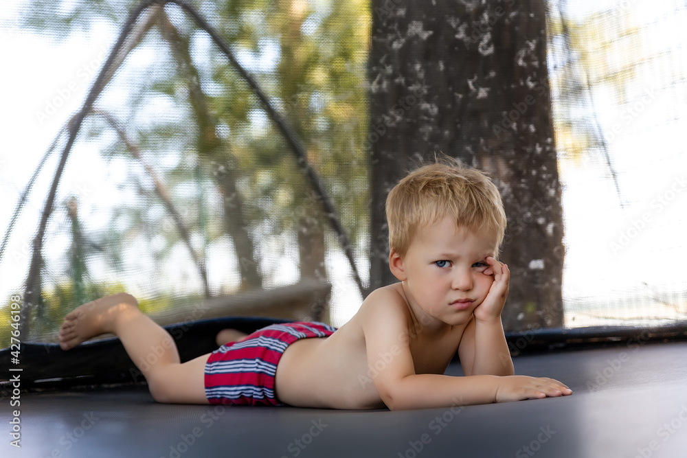 Portrait of cute bored little caucasian funny sad blond toddler boy lying  inside big black trampoline at home backyard playground area outdoors warm  summer sunny day. Children street sport activity Stock Photo |