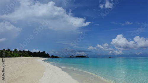 Calm aquamarine ocean and clean sandy beach. There are no people. In the distance, tropical vegetation, a small island. There are picturesque cumulus clouds in the azure sky. Maldives. Endless summer
