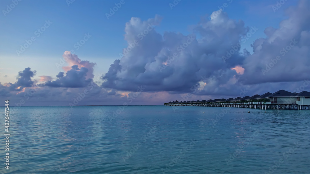 The aquamarine ocean is calm. Above the water - a row of villas. There are scenic blue and pink cumulus clouds in the morning sky. Maldivian idyll.
