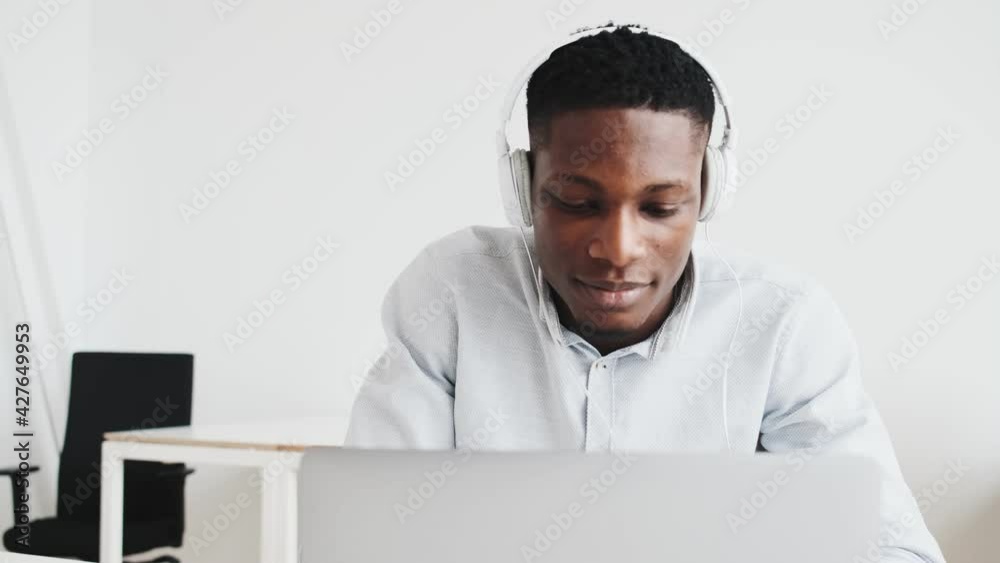A happy black guy with headphones is doing winner gesture using his laptop while sitting in the office A happy black guy with headphones is doing winner gesture using his laptop while sitting in the office