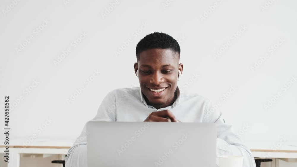 A pleased black guy with earbuds is using a video connection on his laptop sitting alone in the office A pleased black guy with earbuds is using a video connection on his laptop sitting alone in the office