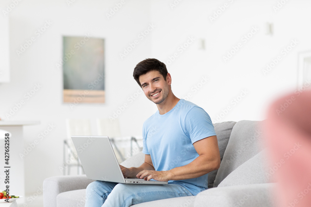 Young smiling man in blue t-shirt with laptop in living room, freelancer
