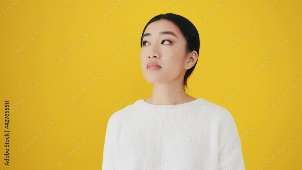 A sad disappointed asian woman is looking to the camera standing isolated over yellow wall in the studio A sad disappointed asian woman is looking to the camera standing isolated over yellow wall in the studio