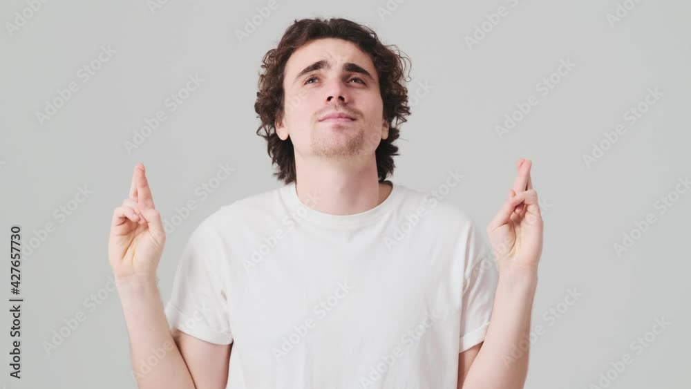 The inspired man with closed eyes praying with crossing fingers and then smilingly looking at the camera while standing in the studio The inspired man with closed eyes praying with crossing fingers and then smilingly looking at the camera while standing in the studio