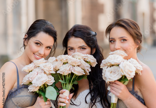 Close up of brunette bride together with bridesmaids bury their faces behind wedding bouquets and look at the camera. Concept of bride whith bridesmaids