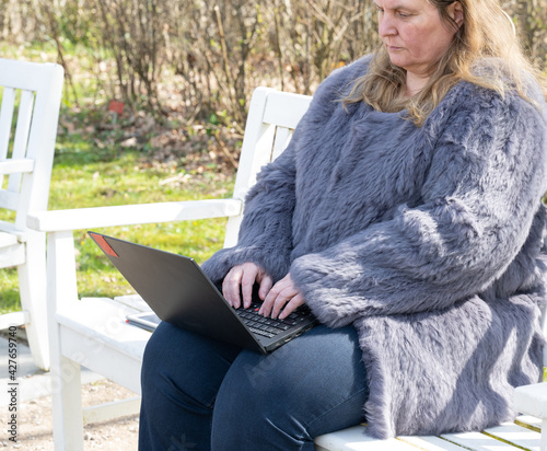 Frau in einer Kaninchenfell Jacke arbeitet im Garten mit einem Notebook im Homeoffice  