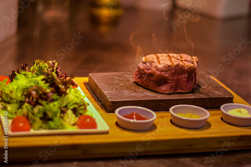 Grilled juicy beef steak smoking on hot stone served on wooden board with salad and sauces