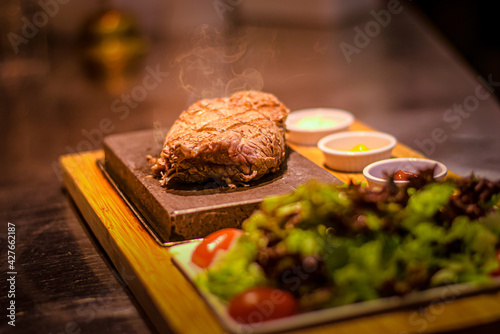 Grilled juicy beef steak smoking on hot stone served on wooden board with salad and sauces