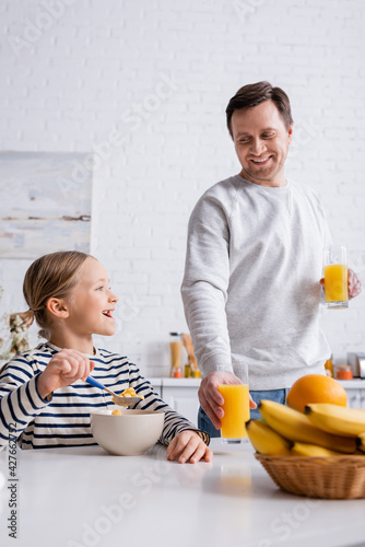 smiling man holding orange juice near daughter eating corn flakes for breakfast