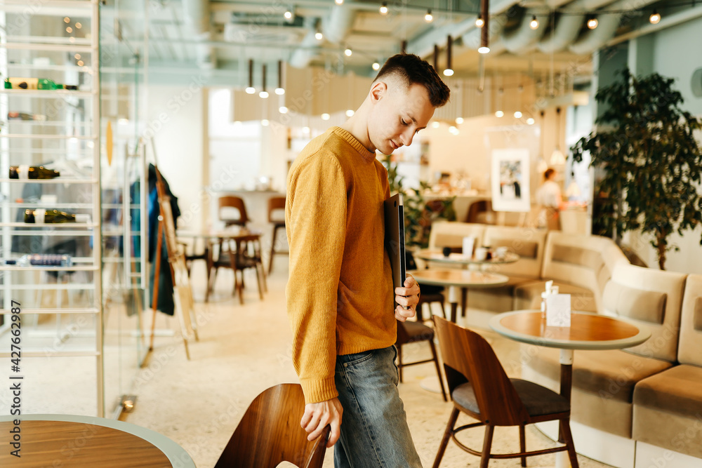 Young smiling business man freelancer in casual yellow sweater and jeans working remotely using laptop and phone sitting in cafe, selective focus Young smiling business man freelancer in casual yellow sweater and jeans working remotely using laptop and phone sitting in cafe, selective focus