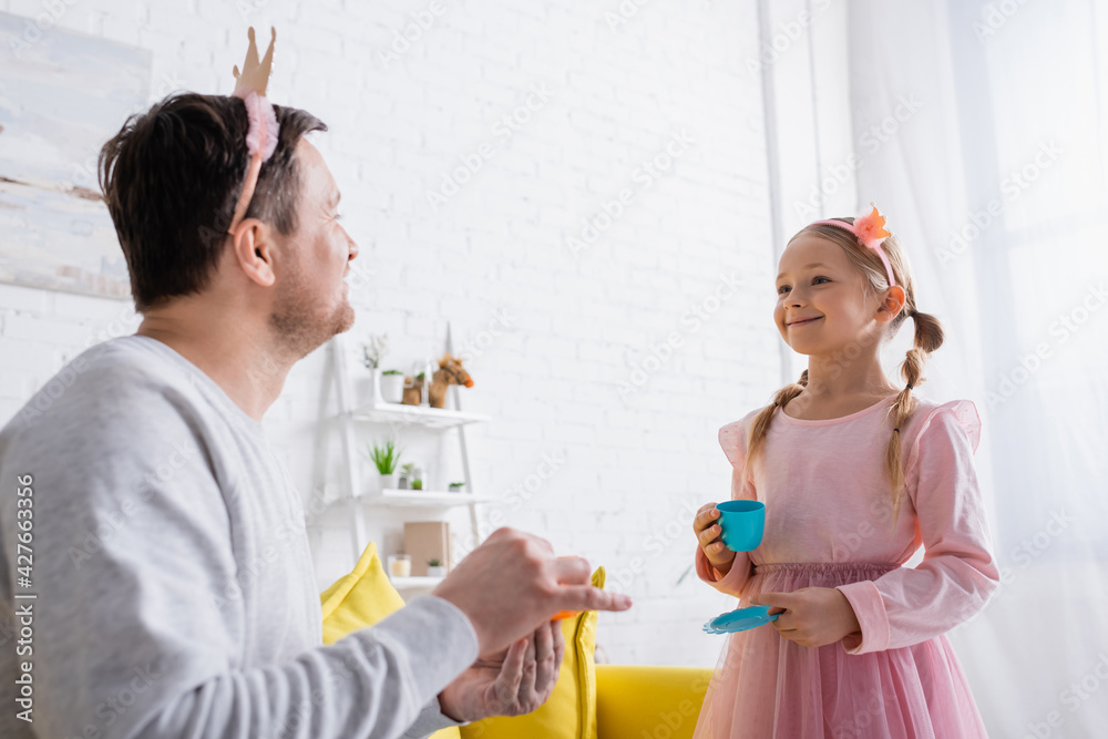 cheerful girl in toy crown looking at father on blurred foreground cheerful girl in toy crown looking at father on blurred foreground