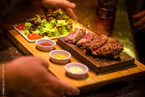 chef service of freshly cut piece of steak on the hot stone with sauces and side salad