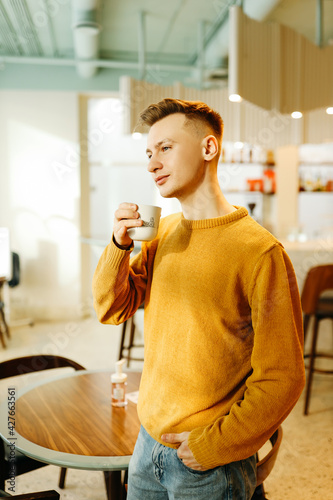 A smiling happy young male student in a bright yellow sweater holds a mug drinks coffee and looks thoughtfully to the side in the indoors cafe