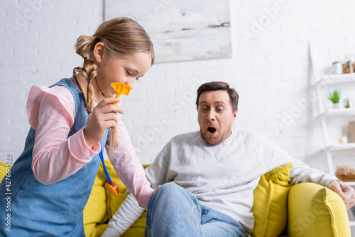 girl holding toy reflex hammer near knee of father sitting on couch with open mouth, blurred background