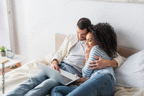 cheerful interracial couple chilling on bed and watching comedy movie on laptop in bedroom