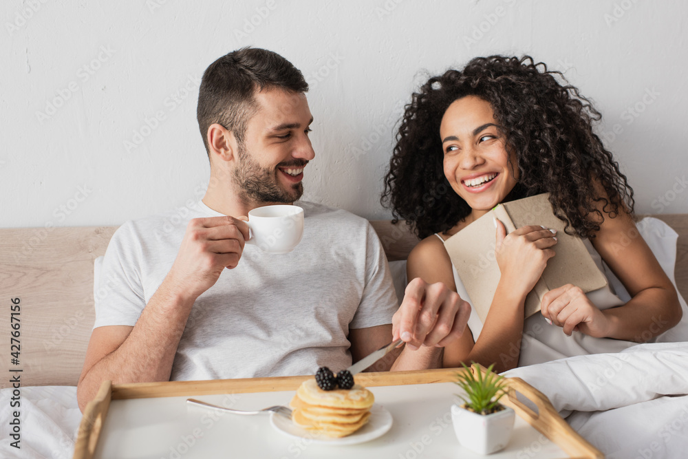 cheerful african american holding book near happy boyfriend having breakfast in bed cheerful african american holding book near happy boyfriend having breakfast in bed
