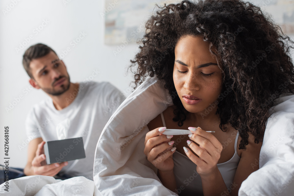 ill african american woman looking at digital thermometer near worried boyfriend on blurred background