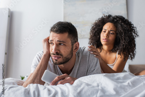 upset man lying on bed and holding smartphone near worried african american woman on blurred background