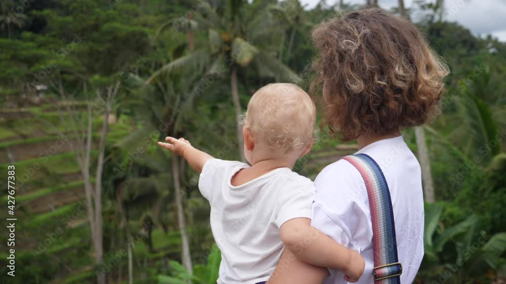 Back view of a Caucasian woman holding her baby looking at the jungle landscape.Blonde baby pointing far away Back view of a Caucasian woman holding her baby looking at the jungle landscape.Blonde baby pointing far away