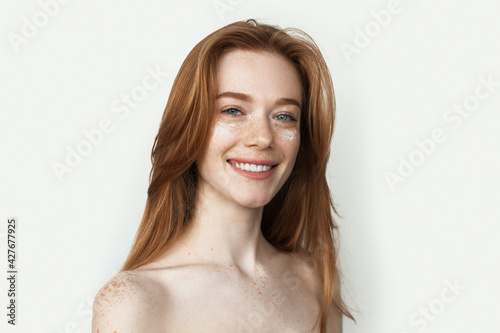 Ginger woman with freckles is massaging her face with a cream smiling with bare shoulders