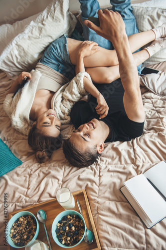 Top view photo of a caucasian couple lying in bed making a selfie while reading a book and eating cereals