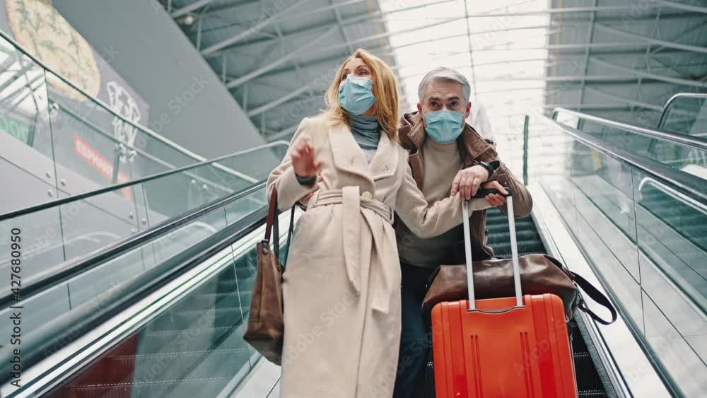 Happy couple of travellers in protective masks going down on escalator with luggage, waving hand to people Happy couple of travellers in protective masks going down on escalator with luggage, waving hand to people