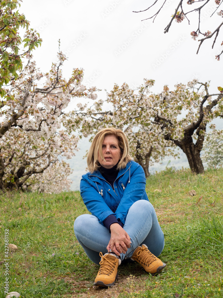 Mature woman seated on the grass in landscape of fields with cherry trees in flowering season Mature woman seated on the grass in landscape of fields with cherry trees in flowering season