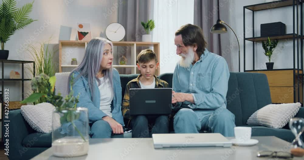 Attractive smiling smart teen boy sitting on the couch between his positive old grandparent and working on laptop while they talking with each other Attractive smiling smart teen boy sitting on the couch between his positive old grandparent and working on laptop while they talking with each other