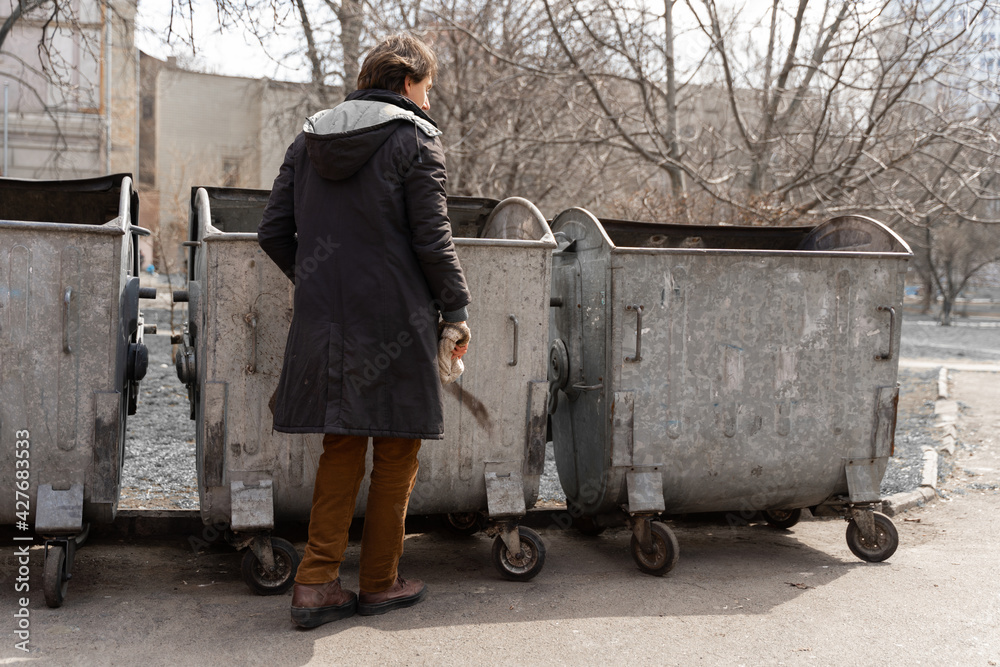 a poor caucasian man is looking for food and valuables in a trash bin on the street. the problem of homeless and unemployed people.