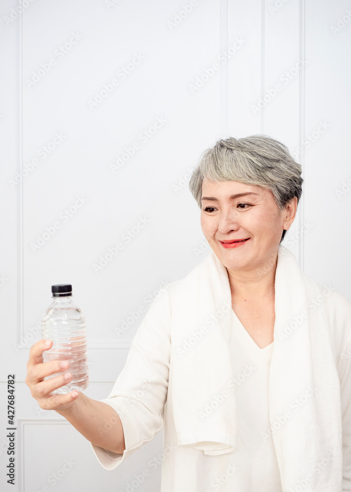 Senior asian woman taking a break while exercising at home. Athletic mature woman holding bottle of water. Senior asian woman taking a break while exercising at home. Athletic mature woman holding bottle of water.