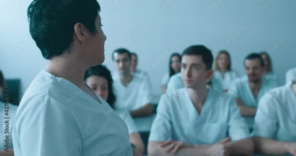 In the lecture hall, a female medical worker is walking and explaining something in front of the male and female medical workers who are sitting at the desk and listening to her speech and take notes. In the lecture hall, a female medical worker is walking and explaining something in front of the male and female medical workers who are sitting at the desk and listening to her speech and take notes.