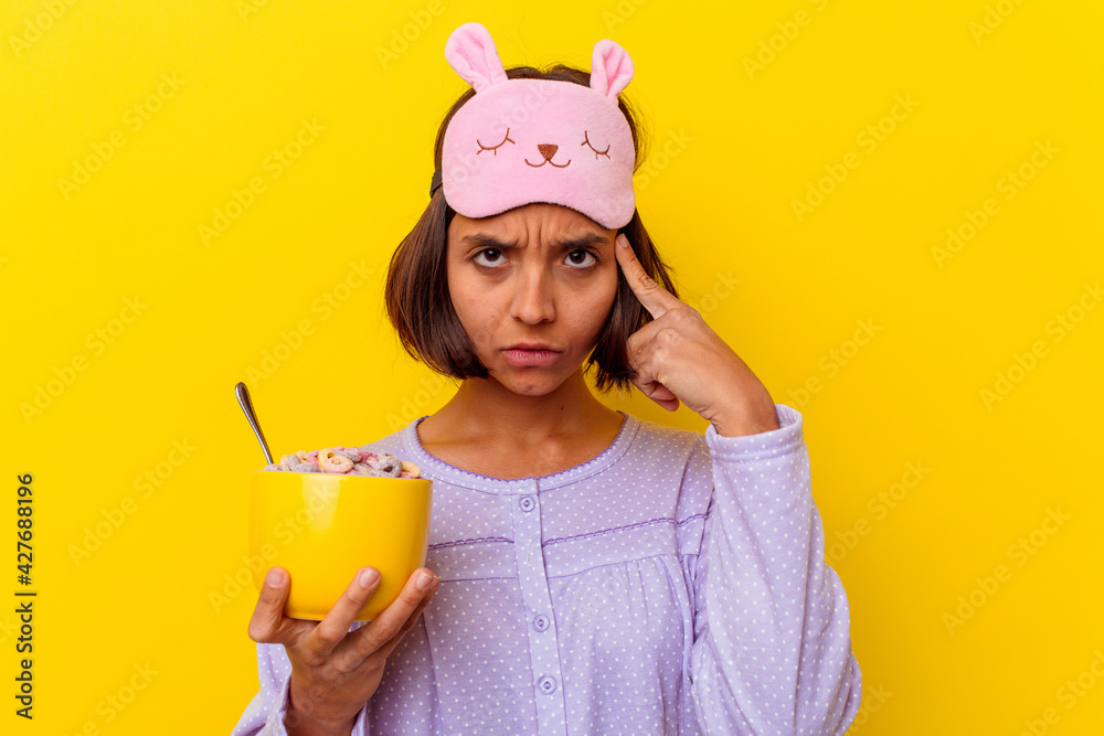 Young mixed race woman eating cereals wearing a pijama isolated on yellow background pointing temple with finger, thinking, focused on a task.