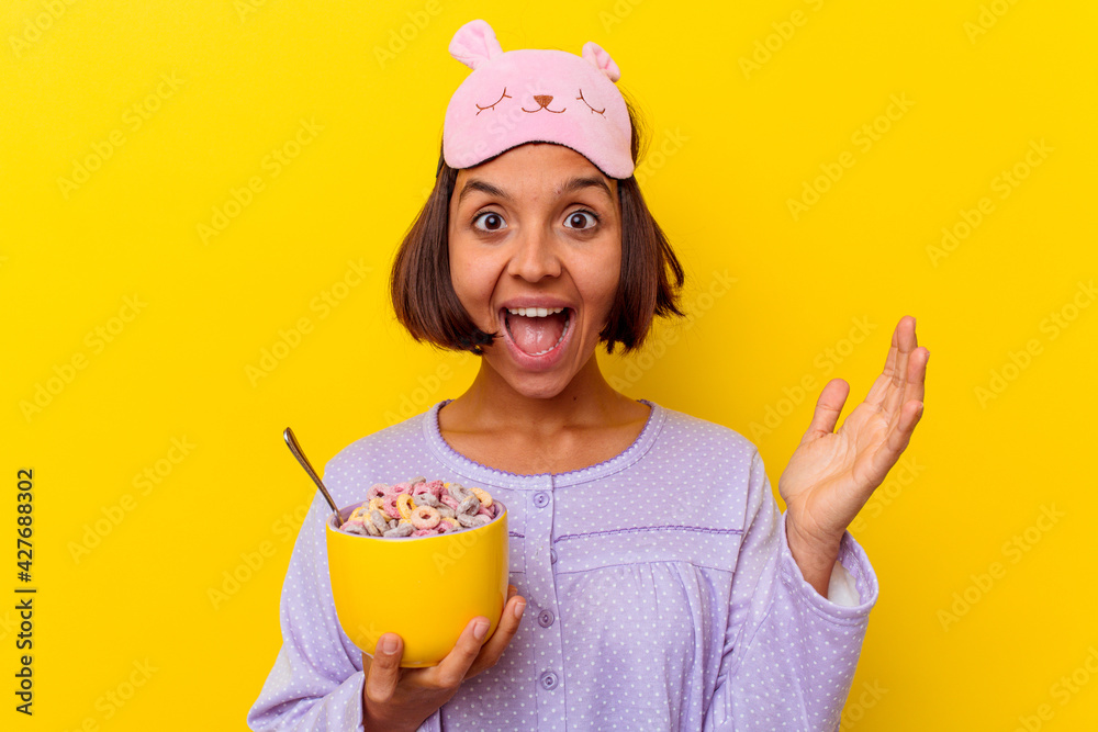 Young mixed race woman eating cereals wearing a pijama isolated on yellow background receiving a pleasant surprise, excited and raising hands. Young mixed race woman eating cereals wearing a pijama isolated on yellow background receiving a pleasant surprise, excited and raising hands.