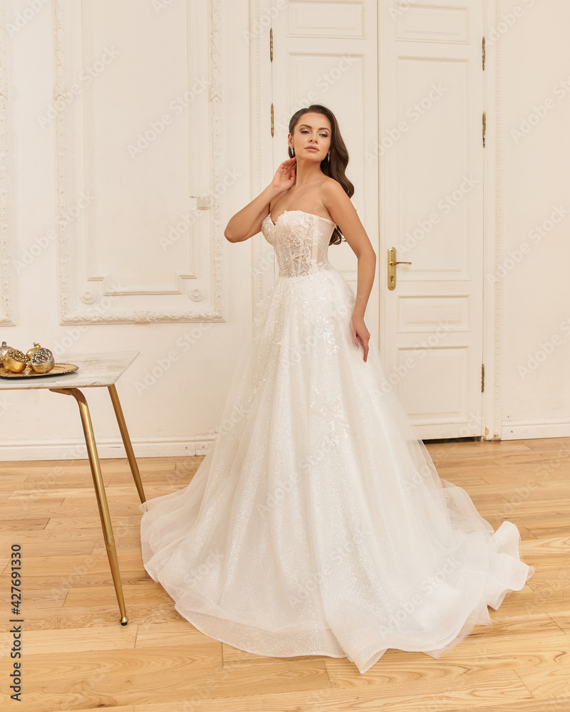 Elegant caucasian woman with natural make-up and hairstyle wearing white bridel dress, standing and posing in bright stylish interior. Full length portrait Elegant caucasian woman with natural make-up and hairstyle wearing white bridel dress, standing and posing in bright stylish interior. Full length portrait