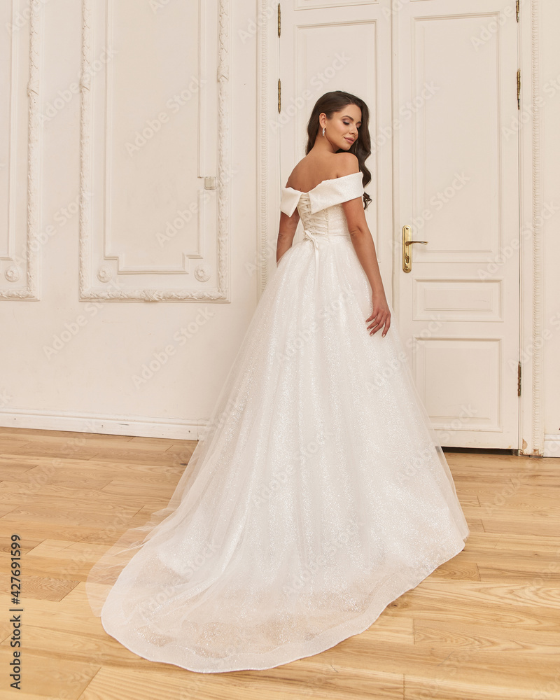 Fototapeta premium Young beauiful happy bride wearing white wedding dress and posing in bright empty interior