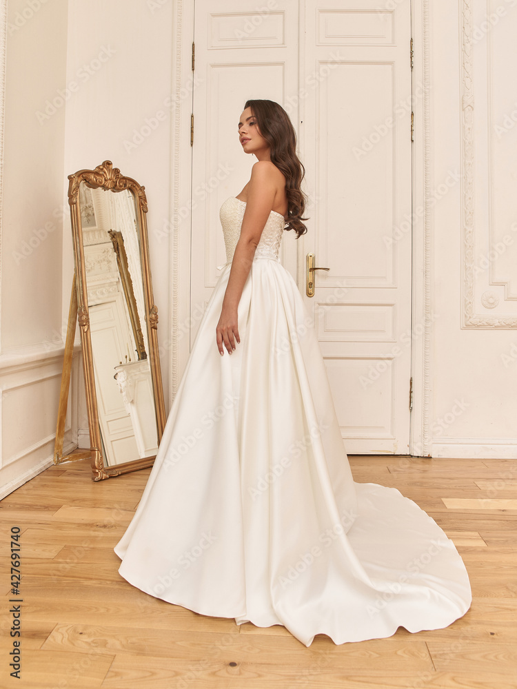 Young elegant bride in white luxury wedding dress standing and posing in bright stylish interior with old mirror Young elegant bride in white luxury wedding dress standing and posing in bright stylish interior with old mirror